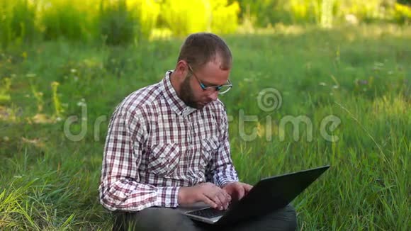 戴眼镜的年轻人坐在草地上在膝上打字视频的预览图