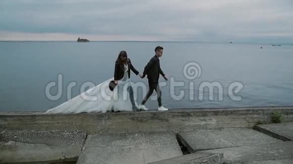 美丽时尚的新娘和新郎在黄昏时沿着堤岸散步慢动作视频的预览图