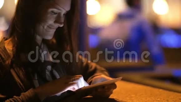 一个漂亮的年轻女人晚上用她的平板电脑她微笑着自然地抚摸着她的头发慢节奏视频的预览图