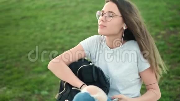 美丽的黑发女孩戴着眼镜坐在公园的草地上从背包里拿出润唇膏滋润皮肤视频的预览图