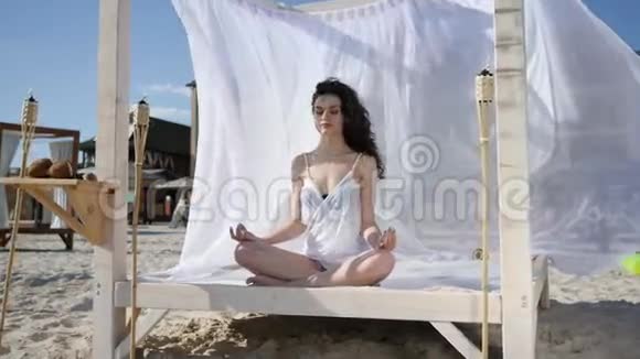 女性在莲花位置冥想在平房瑜伽在海滩在夏天风发展头发和白布热带视频的预览图
