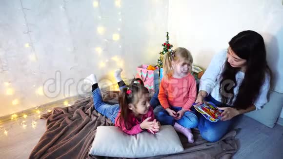美丽的姐姐在圣诞树和圣诞树的背景下和坐在地毯上的小女孩玩耍视频的预览图