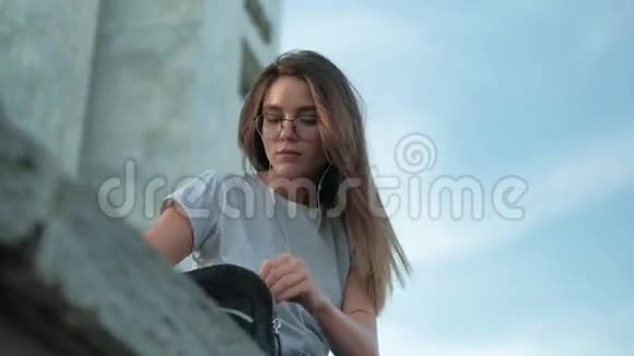 戴眼镜的布鲁内特女孩坐在日落时分听音乐在背包里寻找一些东西视频的预览图