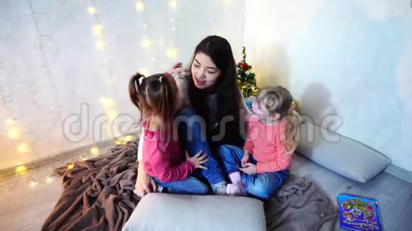美丽的姐姐在圣诞树和圣诞树的背景下和坐在地毯上的小女孩玩耍视频的预览图