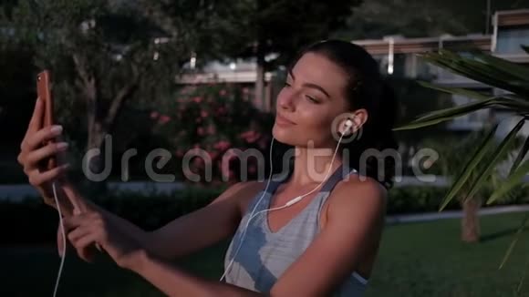 美丽的女人带着耳机在公园前点击电话视频的预览图