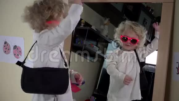 可爱的学步女孩带着粉红色太阳镜手提袋在镜子前摆姿势视频的预览图