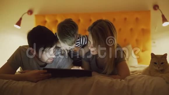 幸福的家庭有一个小儿子和一只有趣的猫躺在家里的床上以前用平板电脑玩游戏视频的预览图