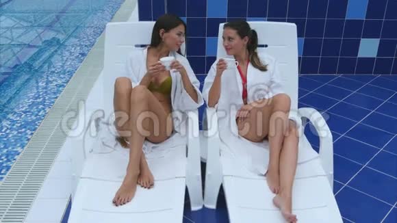 两位微笑的年轻女士坐在泳池边的躺椅上喝茶视频的预览图