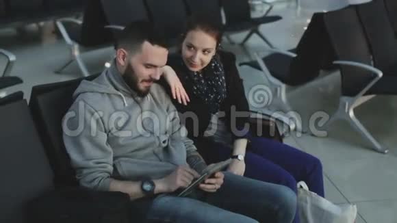 那家伙和穿着休闲服装的女孩看着坐在机场候机大厅的黑色椅子上的平板电脑视频的预览图