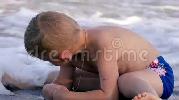 男孩坐在海滩上享受着和沙子一起玩的海浪慢动作视频的预览图