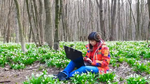 一个漂亮的小女孩正在春天的森林里拿着一台笔记本电脑工作里面满是盛开的雪花视频的预览图