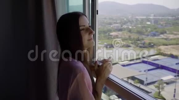 穿着粉红色透明礼服的女人站在那里望着全景的窗户喝着咖啡4k华欣视频的预览图