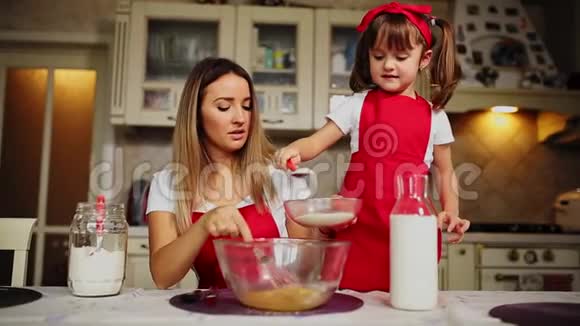 美丽的年轻母亲帮助她的小女儿一起用红色围裙做蛋糕把面粉倒入碗里打一打视频的预览图