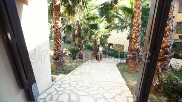 年轻的黑发女人穿着短裤太阳镜和高跟鞋带着手提箱在酒店的棕榈树之间假期视频的预览图