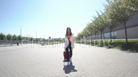 在阳光明媚的日子里穿红衬衫的漂亮女人带着手提箱走在外面视频的预览图