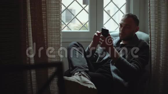 年轻的帅哥坐在客厅的扶手椅上使用智能手机有魅力的商人浏览互联网视频的预览图