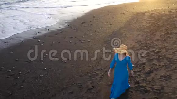 鸟瞰一个女孩穿着蓝色连衣裙在海滩上散步日落时有黑色的沙子西班牙加那利群岛特内里费视频的预览图