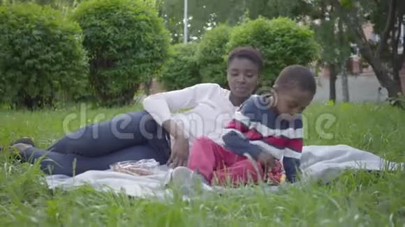 漂亮的非裔美国女人和她的小儿子躺在公园的毯子上可爱的孩子花时间视频的预览图