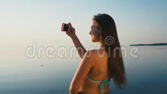 美丽的年轻女子在海边海滩的日落时用智能手机拍照度假慢动作视频的预览图