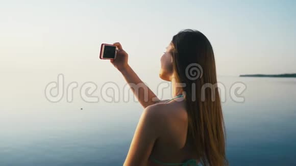 美丽的年轻女子在海边海滩的日落时用智能手机拍照度假慢动作视频的预览图