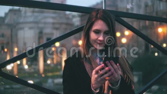 年轻漂亮的女人晚上坐在城市里使用智能手机女孩用触摸屏键盘发短信视频的预览图