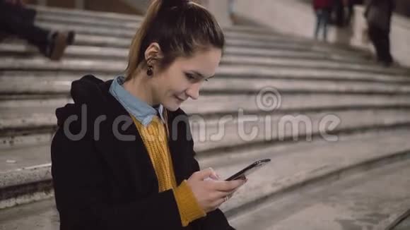年轻女子坐在梯子上拿着智能手机的肖像女孩使用触摸屏浏览互联网视频的预览图