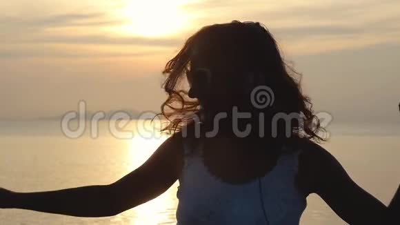 年轻的黑发快乐的女人在美丽的日落海滩上用耳机听音乐玩得很开心慢动作视频的预览图