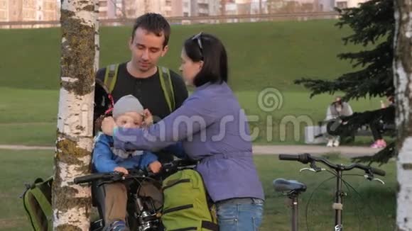 家长在公园里给幼儿戴上自行车头盔一个男孩坐在自行车前的儿童座位上视频的预览图