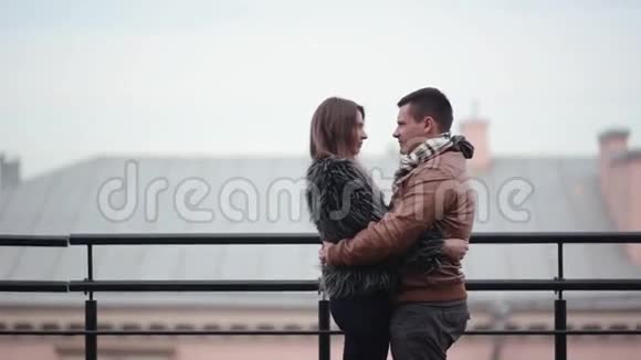 可爱的夫妇在寒冷的天气里站在屋顶上亲吻和大笑他们互相拥抱穿着毛皮外套的女孩侧视视频的预览图