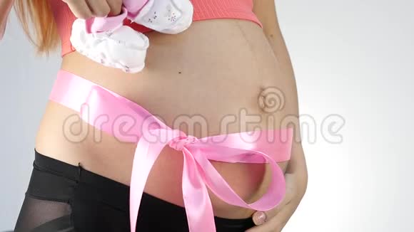 穿着内衣的怀孕妈妈在玩婴儿鞋腹部有粉红色的丝带慢动作视频的预览图