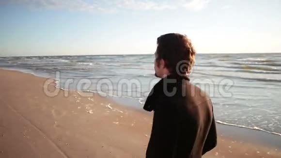 穿着黑色外套的年轻人走在海岸边太阳在他的背上闪闪发光视频的预览图