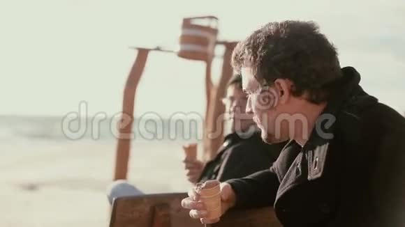 两个年轻人坐在岸边的一张木凳上吃着冰激凌聊天视频的预览图