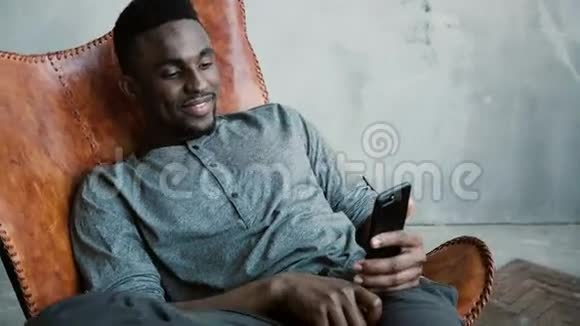 非洲男子坐在椅子上的肖像使用智能手机人读留言笑打字按摩微笑着视频的预览图