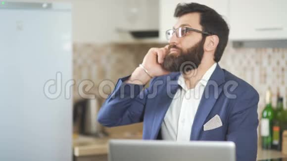 戴眼镜留胡子的人在家庭办公室工作手机和笔记本电脑视频的预览图