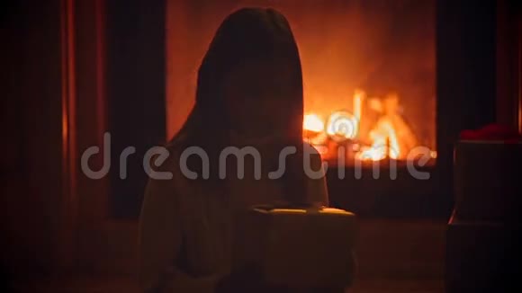 可爱的女孩坐在黑暗的客厅里在燃烧的壁炉和打开的盒子与圣诞礼物视频的预览图