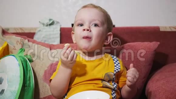一个孩子坐在家里的沙发上看电视吃玉米棒微笑视频的预览图