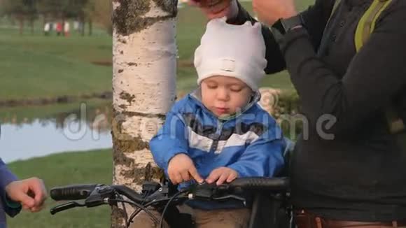 家长在公园里给幼儿戴上自行车头盔一个男孩坐在自行车前的儿童座位上视频的预览图