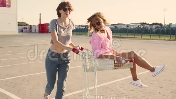 时尚有趣的年轻时髦女孩在购物中心停车场玩得很开心在街上的购物车里互相滚动慢动作视频的预览图
