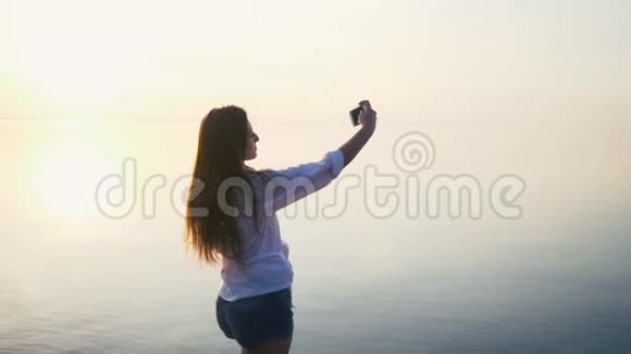 美丽的年轻女子在日落时用她的智能手机在海边度假慢动作视频的预览图