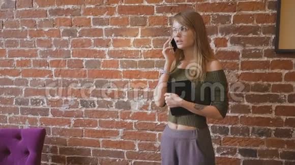 戴眼镜的年轻可爱的女人站在靠近砖墙的房间里拿着平板电脑一边用手机说话视频的预览图