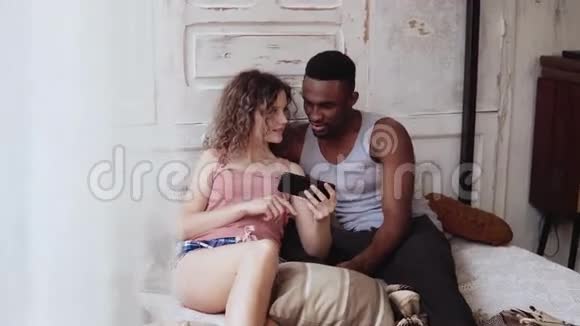 多种族夫妇穿着睡衣坐在床上使用智能手机非洲男人和欧洲女人上网微笑视频的预览图