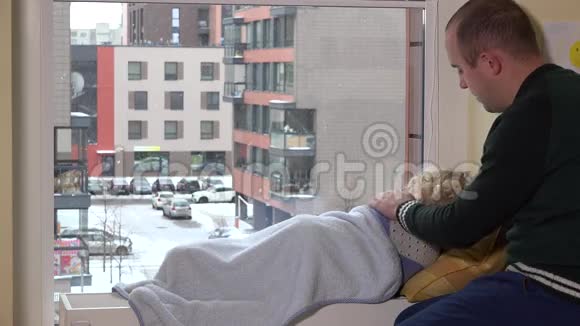 小心的父亲抚摸他躺在窗户附近散热器上的孩子下雪了视频的预览图