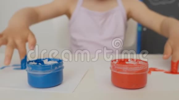 一个小可爱的女孩坐在桌子旁用明亮的手指颜料画纸把她的手指浸在颜料罐里视频的预览图