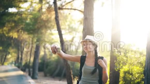 有趣的戴帽子的旅游女孩在旅行和搭便车期间用智能手机相机自拍照片视频的预览图