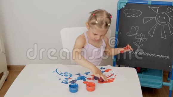 一个小可爱的女孩坐在桌子旁用明亮的手指颜料画纸把她的手指浸在颜料罐里视频的预览图
