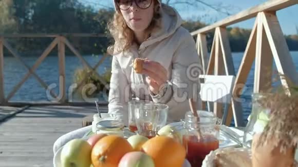 年轻女子穿着暖和的衣服吃着汉堡在木桥边河边野餐周末寒冷的天气野营视频的预览图