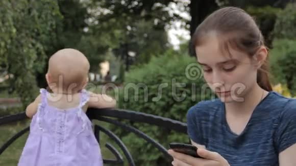一个非常年轻的保姆一边接着智能手机一边说话一边婴儿站在她旁边背着长凳视频的预览图