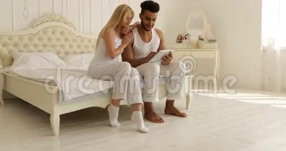 双人坐床使用平板电脑混合种族男子女子微笑早间卧室视频的预览图