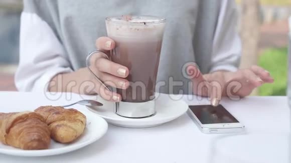 年轻女子在智能手机上写信息同时喝可可和牛角面包女性在早晨使用手机视频的预览图