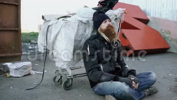 寒冷的冬天坐在购物车旁的人行道上的一个留着胡须的流浪汉和一个垃圾容器视频的预览图